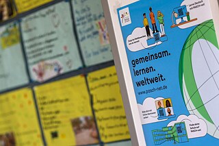 PASCH-net-Poster mit verschiedenen Grafiken (Weltkugel, Personen, Schulgebäude) an einer Pinnwand in einem Klassenzimmer