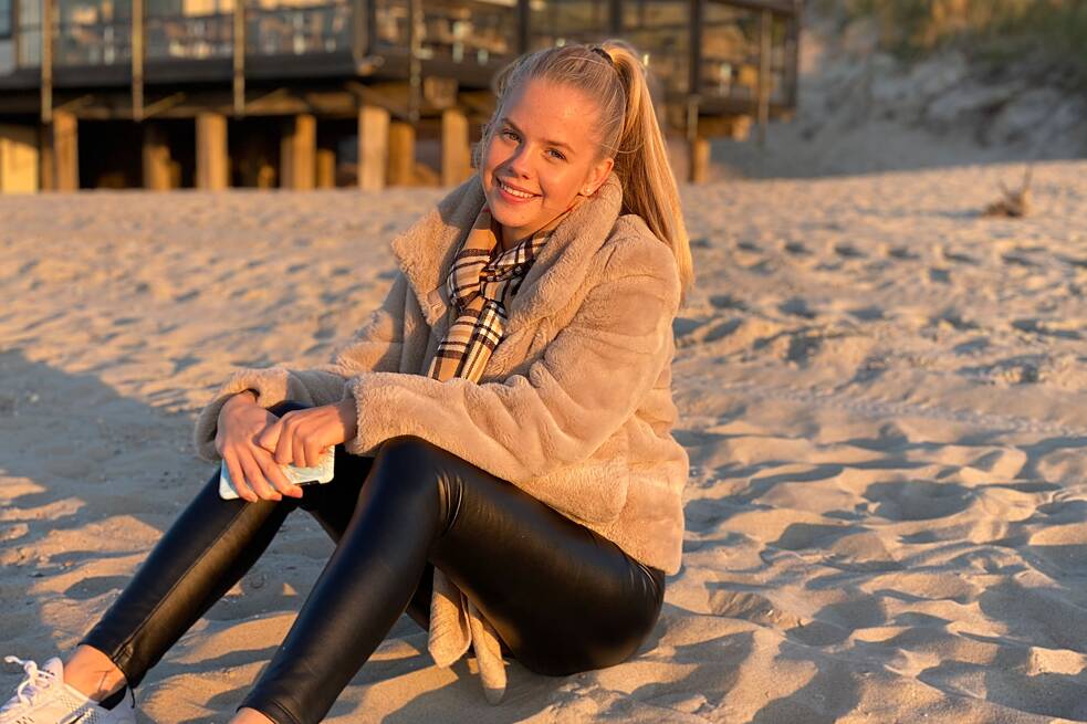 Jugendliche in heller Jacke und schwarzer Leggins sitzt im Sand am Strand