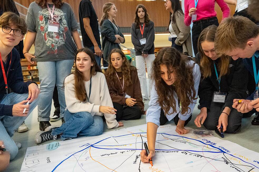 Schülerinnen und Schüler sitzen auf dem Boden und schreiben zusammen auf ein großes, weißes Plakat