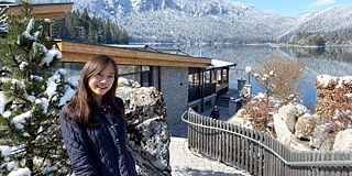 Frau in dunkler Jacke steht vor vor einer Holzhütte an einem verschneiten Gewässer