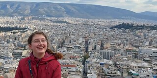 Frau in roter Jacke vor einem Stadtpanorama, im Hintergrund Berge