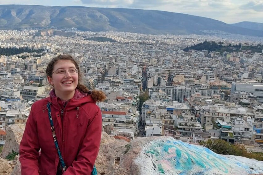 Frau in roter Jacke vor einem Stadtpanorama, im Hintergrund Berge