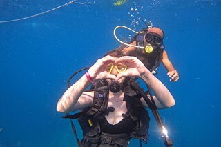 Zu sehen sind zwei Personen in Taucherausrüstung unter Wasser. Beide schauen in die Kamera. Die vordere Person formt mit den Händen ein Herz