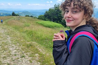 Frau von der Seite aufgenommen, die in der Hand eine kleine lilane Blume hält. Im Hintergrund ist eine hügelige, grüne Landschaft und vereinzelte Personen zu sehen.